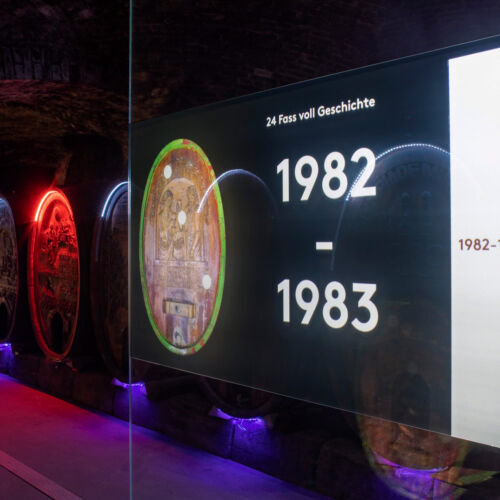 Ein Foto des Touchmonitors „Fassgang" in der Kellergenossen-Ausstellung. Im Hintergrund leuchten Weinfässer.
